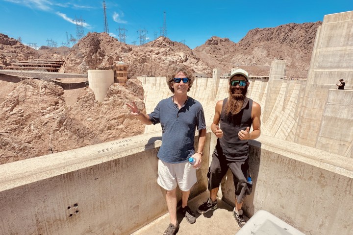 a man standing next to a brick building with a mountain in the background