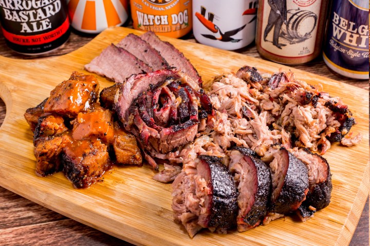 Assorted barbecued meats on a wooden board with beer bottles in the background.