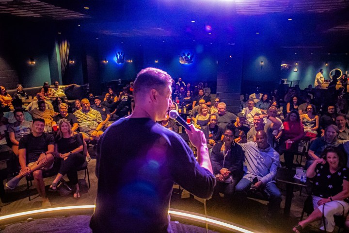 Person performing stand-up comedy on stage with audience laughing in a dimly lit comedy club.