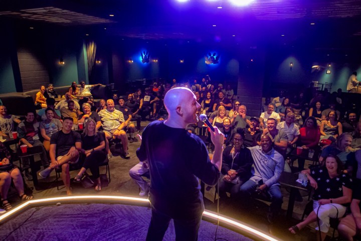 Comedian on stage performing to a laughing audience in a dimly lit comedy club.