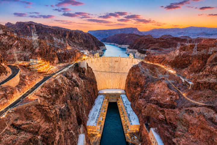 Aerial view of a large dam with a river, rugged cliffs, and a colorful sunset sky.